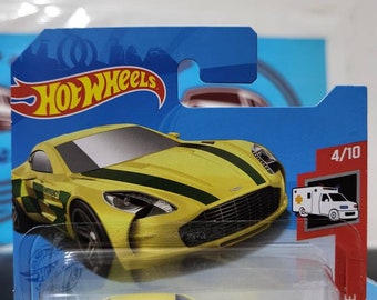 May include: A yellow Hot Wheels die-cast toy car with green and yellow stripes and the word "EMERGENCY" on the side. The car is in a plastic package with a blue background and the Hot Wheels logo. The package also includes a small image of an ambulance.