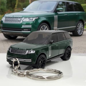 May include: A dark green Range Rover toy car with black wheels and a silver keychain ring. A full-size dark green Range Rover is in the background. The toy car has the words "Range Rover" on the front.