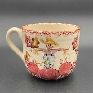 May include: Cream-colored ceramic mug with a handle, featuring a hand-painted harvest design. The design includes a scarecrow, pumpkins, and fall foliage in shades of red, orange, and yellow. The rim has a decorative border.