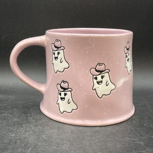 May include: A pink ceramic mug featuring white ghost illustrations wearing cowboy hats. The mug has a large handle and a speckled finish. The ghosts have smiling faces and are outlined in black.
