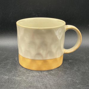 May include: A ceramic mug with a textured, light gray body and a gold band around the bottom. The mug has a rounded handle.