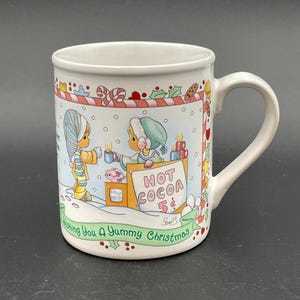 May include: White ceramic mug with a Christmas-themed illustration. The design features two cartoon figures selling hot cocoa for 5 cents. The mug is decorated with candy canes, holly, and the text "Wishing You A Yummy Christmas."