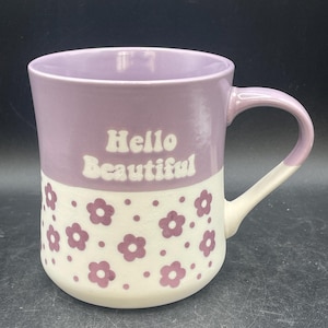 May include: A ceramic mug with a lavender top and a white bottom. The mug has a handle and is decorated with purple flower patterns. The words "Hello Beautiful" are printed in white on the mug.