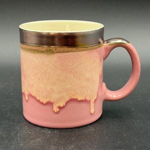 May include: A ceramic mug featuring a pink base with a cream-colored glaze dripping down from the top. The upper rim and handle are finished in a metallic bronze glaze. The interior of the mug is a solid cream color, and the handle is pink.