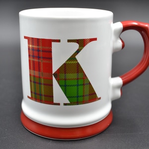 May include: White ceramic mug with a red handle and base. The letter "K" is prominently displayed on the front, filled with a red, green, and gold plaid pattern. The mug is designed for hot beverages.