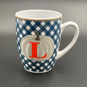 May include: White ceramic mug with a blue and white checkered pattern. The mug features a gray pumpkin with the red letter "L" on the front. The rim has a gold trim and a white handle.
