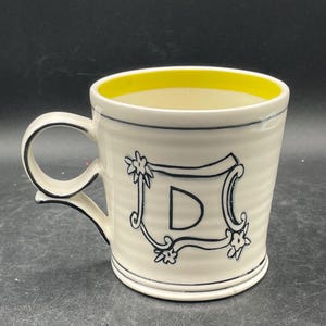 May include: Cream-colored ceramic mug with a yellow interior and a black handle. The mug features a black outlined letter "D" within a decorative frame with floral accents. The rim and base have black trim.
