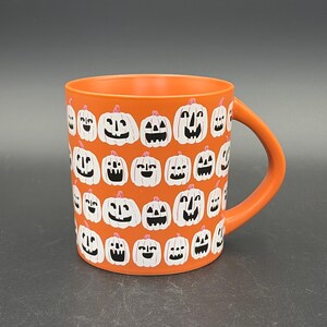 May include: A bright orange ceramic mug featuring a repeating pattern of white jack-o'-lanterns with black carved faces. The mug has a smooth matte finish, a cylindrical body, and a curved handle on the right side, showcasing a festive seasonal design.