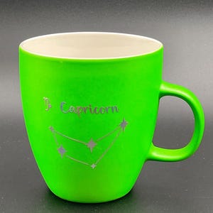 May include: A bright green ceramic mug with a white interior. The mug features the word "Capricorn" and a constellation design in silver. The mug has a curved handle and a smooth, glossy finish.