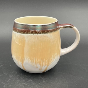 May include: A ceramic mug with a rounded body and a handle. The mug features a gradient of colors, starting with a cream-colored interior and transitioning to a light orange exterior. The rim has a dark brown band with a green iridescent sheen.