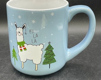 May include: A light blue ceramic mug with a white interior. The mug features a cartoon llama wearing a green and red scarf, a Christmas tree, and the text "FA LA LA". Snowflakes are scattered around the design.