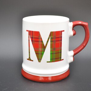 May include: White ceramic mug with a red base and handle. The letter "M" is prominently displayed on the front in a red and green plaid pattern. The mug has a classic, festive design.