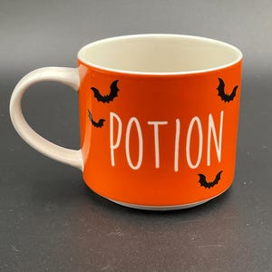 May include: Orange ceramic mug with the word "POTION" in white lettering and black bat silhouettes. The mug has a white handle and interior, perfect for Halloween or spooky season.