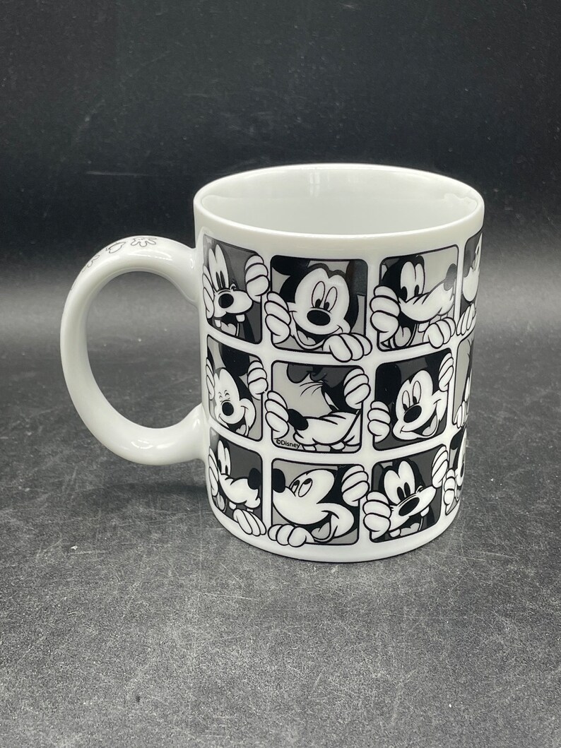 Pu&ograve; includere: Tazza in ceramica bianca con un design bianco e nero di Topolino che spunta da quadrati. La tazza ha un manico nero.