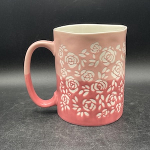 May include: A ceramic mug with a gradient of pink hues, adorned with a repeating white rose pattern. The handle and interior are a solid pink, creating a contrast with the floral design. The mug is suitable for hot beverages.