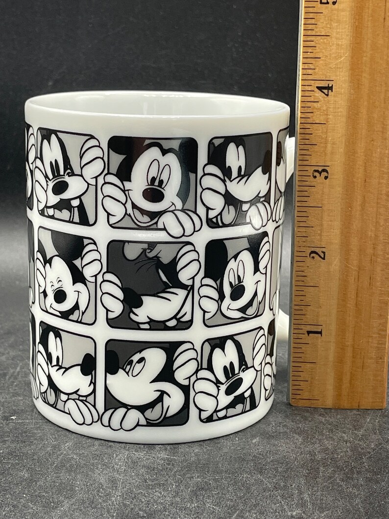 Pu&ograve; includere: Tazza in ceramica bianca con un design bianco e nero di volti di Topolino in quadrati.