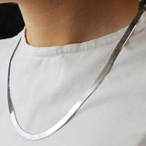 Puede incluir: Un collar de cadena de espiga plateada. El collar es plano y flexible, con una superficie lisa y reflectante. La cadena est&aacute; drapeada alrededor del cuello, descansando sobre una camiseta blanca.