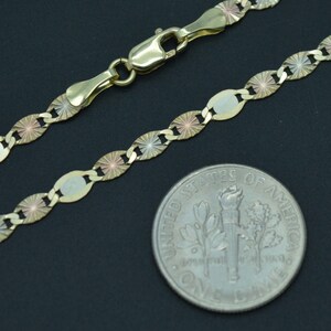 May include: A gold chain necklace with a tri-colour design. The chain is made of oval links with a starburst pattern. The chain is shown next to a US dime for size comparison.