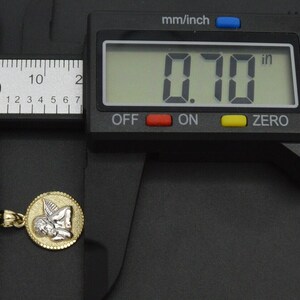 May include: A gold and white angel pendant charm, measuring 0.70 inches in diameter, is shown next to a digital caliper.