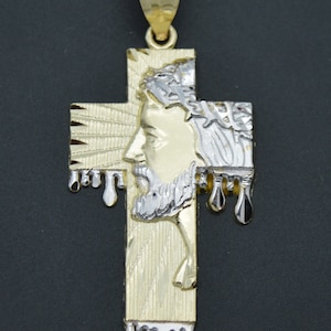 Può includere: Un ciondolo a forma di croce di Gesù in oro e argento bicolore con un'immagine dettagliata del volto di Gesù e una corona di spine. La croce ha un effetto gocciolante, con piccole gocce di metallo che pendono dal basso.