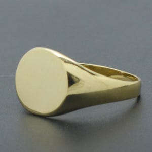 May include: A polished gold signet ring featuring a smooth, flat, circular face and a solid, tapered band. The metal surface is reflective and uniform in colour, showcasing a classic, simple design with no engravings or additional decorative elements.