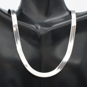 May include: A silver-colored herringbone chain necklace displayed on a black mannequin. The necklace has a flat, wide design with a reflective surface. The chain is draped in a gentle curve, showcasing its sleek and modern style.