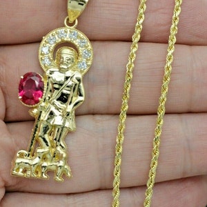 May include: Gold chain necklace with a pendant featuring a religious figure with a red gemstone and clear stones. The pendant is detailed with a halo and a staff.