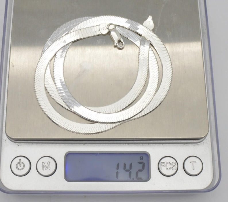 Puede incluir: Un collar de cadena de espiga plateada con cierre de mosquet&oacute;n, enrollado sobre una balanza digital. La balanza muestra un peso de 14,2. El collar est&aacute; hecho de eslabones planos e interconectados.