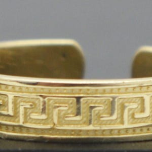 May include: A gold-coloured open band ring featuring a continuous embossed Greek key pattern around the exterior. The metal surface has a polished finish with textured detailing within the geometric meander design, and the band is open at the back.
