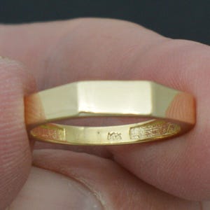 May include: A gold ring with a rectangular top and a faceted design. The ring is held between fingers, showcasing its polished surface and the subtle details of its construction. The ring appears to be a size 8.