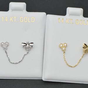 May include: Two pairs of 14K gold earrings on white display cards. One pair is silver-toned with a bee and a cluster of three clear stones, connected by a chain. The other pair is gold-toned with a bee and a cluster of three clear stones.