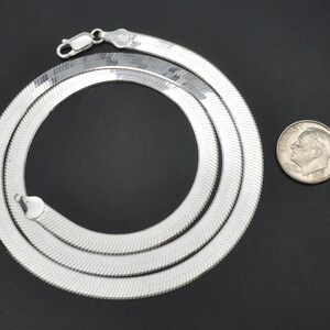 Puede incluir: Un collar de cadena plateada en espiga con cierre de mosquet&oacute;n, dispuesto en forma circular. Una moneda se coloca junto al collar para comparar el tama&ntilde;o. El collar tiene una superficie brillante y reflectante.