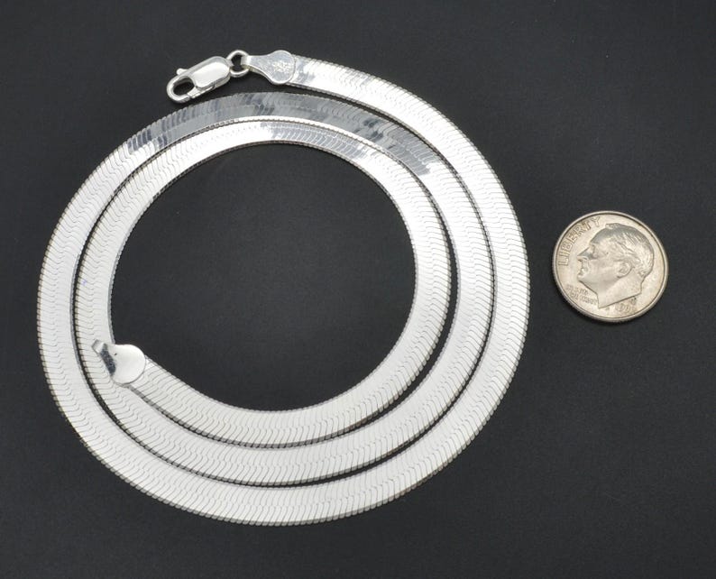 Puede incluir: Un collar de cadena plateada en espiga con cierre de mosquet&oacute;n, dispuesto en forma circular. Una moneda se coloca junto al collar para comparar el tama&ntilde;o. El collar tiene una superficie brillante y reflectante.