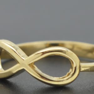 May include: A polished gold ring featuring a prominent infinity symbol as the centerpiece. The band is smooth and circular, connecting to the sides of the horizontal figure-eight design. The metal has a reflective, bright yellow gold finish throughout.