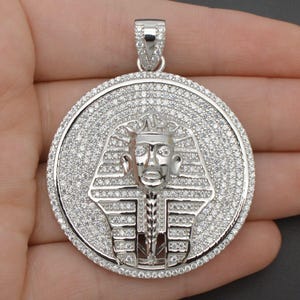 May include: Silver pendant featuring a detailed depiction of an Egyptian pharaoh's head, surrounded by sparkling white cubic zirconia stones.