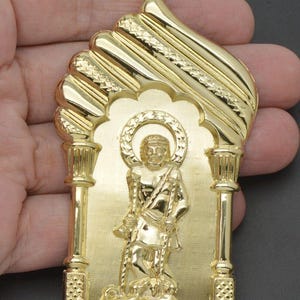 May include: A gold pendant featuring a detailed depiction of a saint holding a chain with two dogs at his feet. The pendant is framed by an ornate archway with a textured design.