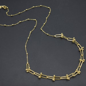May include: A gold-toned necklace featuring a chain with small evenly spaced spherical beads. One section of the necklace transitions into a series of elongated rectangular links, each accented with small gold spheres at the connection points.