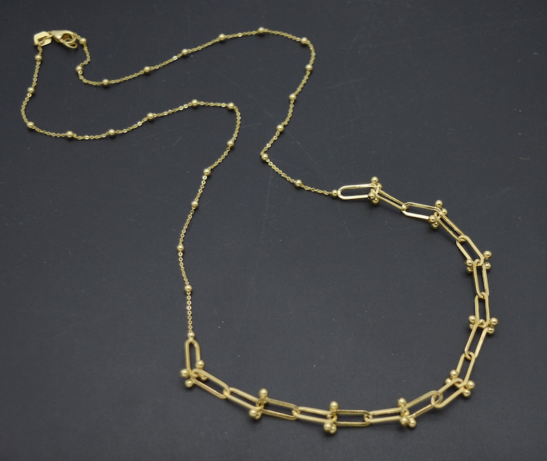 May include: A gold-toned necklace featuring a chain with small evenly spaced spherical beads. One section of the necklace transitions into a series of elongated rectangular links, each accented with small gold spheres at the connection points.