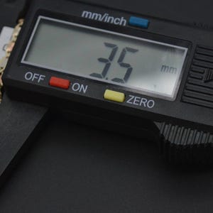 May include: A black digital caliper measuring 3.5 mm. The caliper has a black display with white numbers, red and yellow buttons, and a black and silver handle.