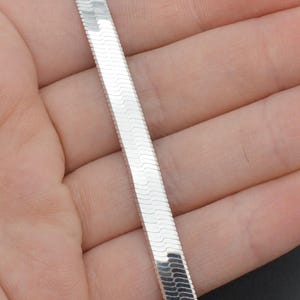 Puede incluir: Un collar de cadena de espiga plateado y brillante. El dise&ntilde;o plano y tejido refleja la luz, creando un efecto brillante. El collar se sostiene en una mano, mostrando su estilo elegante y moderno. Una pieza de joyer&iacute;a cl&aacute;sica.