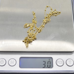 May include: A gold-toned chain necklace featuring a combination of elongated oval links and small spherical beads. The necklace is resting on a digital scale that displays a weight of 3.0 grams, providing a clear measurement of the jewellery piece.