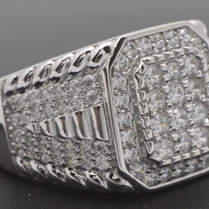 May include: A silver ring with a rectangular face, densely set with small, clear gemstones. The band has a braided pattern on the sides, and the face features a central square of stones surrounded by more gems.