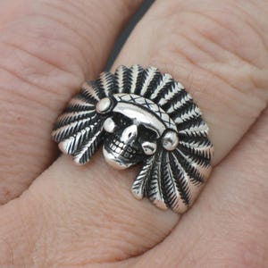 Könnte beinhalten: Ein silberner Ring mit einem Totenkopf, der einen Federschmuck trägt. Der Ring wird an einem Finger getragen.