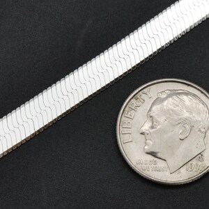 Puede incluir: Un collar de cadena de espiga plateada junto a una moneda. La cadena tiene un dise&ntilde;o plano y tejido. La moneda muestra el perfil de un hombre.