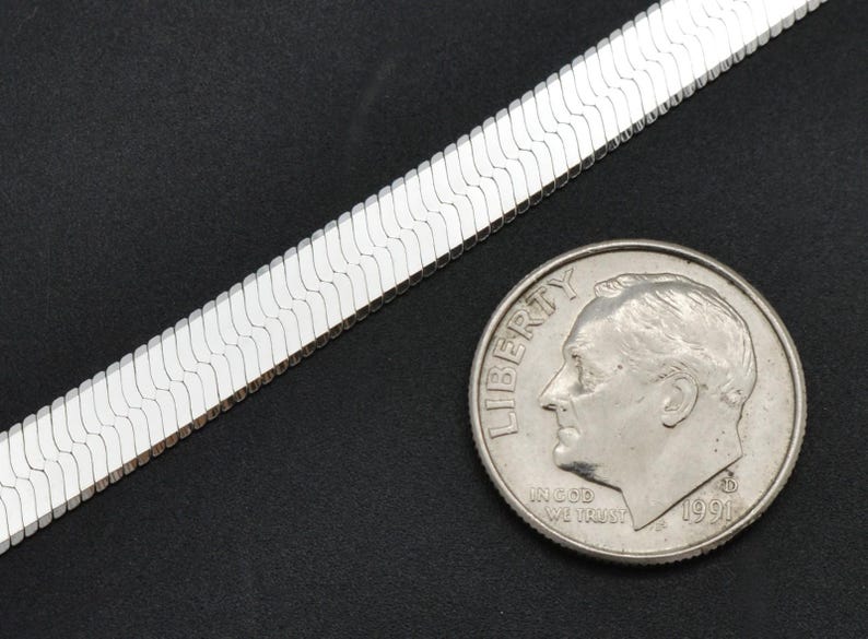 Puede incluir: Un collar de cadena de espiga plateada junto a una moneda. La cadena tiene un dise&ntilde;o plano y tejido. La moneda muestra el perfil de un hombre.