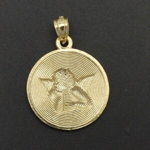 May include: A gold-tone pendant featuring a detailed, engraved image of an angel with wings. The pendant has a circular shape and a textured, concentric circle design.