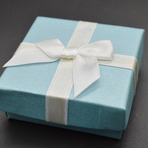 May include: A square light blue gift box with a textured finish, featuring a white satin ribbon tied in a bow across the top. The box has a lid that sits flush with the base, creating a clean and simple presentation for small items or jewellery.