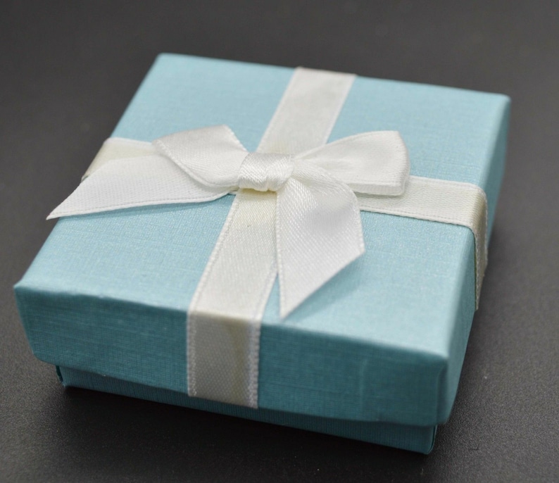 May include: A square light blue gift box with a textured finish, featuring a white satin ribbon tied in a bow across the top. The box has a lid that sits flush with the base, creating a clean and simple presentation for small items or jewellery.