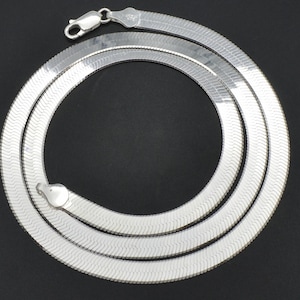 Puede incluir: Un collar de cadena de espiga plateado con un cierre de mosquet&oacute;n. El collar est&aacute; dispuesto en un patr&oacute;n circular sobre un fondo negro. La cadena tiene una superficie plana y lisa con un brillo sutil y reflectante.