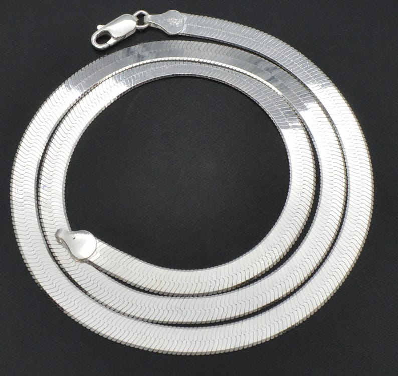 Puede incluir: Un collar de cadena de espiga plateado con un cierre de mosquet&oacute;n. El collar est&aacute; dispuesto en un patr&oacute;n circular sobre un fondo negro. La cadena tiene una superficie plana y lisa con un brillo sutil y reflectante.